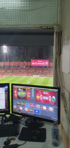 Desde la Cabina de Megafonía: Interpretación Simultánea Búlgaro-Español en el Clasificatorio Mundial 2026 (España vs. Bulgaria) Intérprete búlgaro-español realizando interpretación simultánea desde la cabina de control y megafonía del Estadio José Zorrilla en Valladolid, durante el partido clasificatorio para la Copa Mundial 2026 entre las selecciones de España y Bulgaria. Se aprecian los equipos técnicos de audio, el micrófono y los auriculares, así como una pantalla mostrando la retransmisión del partido con el marcador de "España vs. Bulgaria" y el marcador general del estadio de fondo.